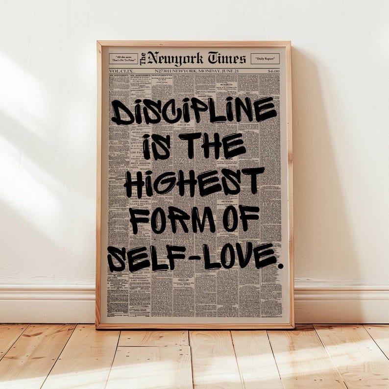 May include: A framed print featuring a vintage New York Times newspaper page. Bold black text reads "DISCIPLINE IS THE HIGHEST FORM OF SELF-LOVE." The print is in a light wooden frame, displayed against a white wall and wooden floor.