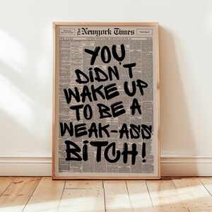 May include: A framed print featuring a newspaper background with bold black text. The text reads "YOU DIDN'T WAKE UP TO BE A WEAK-ASS BITCH!". The print is in a light brown wooden frame, suitable for wall decor.
