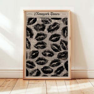 May include: A framed print featuring a pattern of black lipstick kiss marks over a vintage newspaper background. The newspaper headline reads "The New York Times" with the date "Monday, June 21". The print is displayed in a wooden frame.