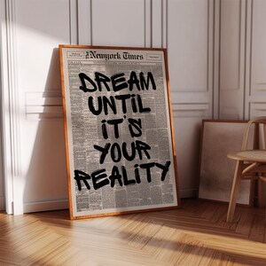 May include: A framed print featuring a newspaper background with the words "DREAM UNTIL IT'S YOUR REALITY" in bold black letters. The frame is a warm brown color, and the print leans against a white wall.