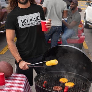 Puede incluir: Una camiseta y gorra negras con la palabra "BELIEVE" impresa. La camiseta también tiene el texto "JOHN 3:16". Una persona sostiene una taza roja y está asando comida en una parrilla de carbón negra. Un mantel a cuadros rojos y blancos está en primer plano.