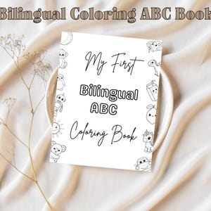 May include: A white coloring book titled "My First Bilingual ABC Coloring Book" with cartoon illustrations of animals and objects. The book is on a white plate, with a sprig of dried flowers to the left.