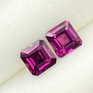 May include: Two square-cut, vibrant magenta gemstones are displayed in a white, foam-lined box. The gems have a faceted surface, catching the light and showcasing their rich color. The box is set against a neutral background.