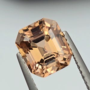 May include: A peach-coloured, rectangular gemstone held by tweezers. The gem has internal reflections. The tweezers are silver and hold the gem against a light background. The gem is cut in an emerald cut.