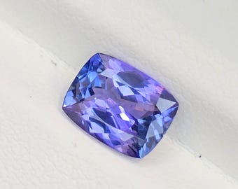 Puede incluir: Una piedra preciosa de tanzanita con corte cojín que presenta una mezcla vibrante de tonos violetas, azules y púrpuras profundos. La piedra tiene una superficie facetada que refleja la luz, mostrando su claridad y transparencia.