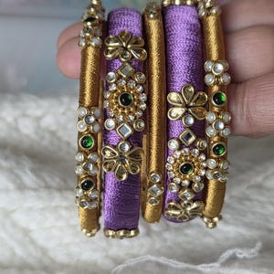 Peut inclure: Ensemble de bracelets ornés. Les bracelets présentent une base dorée avec des motifs floraux et géométriques complexes. Les bracelets centraux sont violets vifs, tandis que les autres sont dorés. Ils sont ornés de pierres précieuses vertes et blanches.