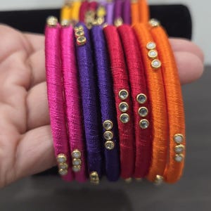 May include: A set of stacked bangles wrapped in vibrant silk thread in shades of hot pink, purple, red, and orange. Each bangle features a small cluster of three clear, round rhinestones set in gold-toned metal accents along the outer surface.