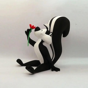 May include: A figurine of Pepe Le Pew, a cartoon skunk, in a seated position. The skunk is black and white, holding a red flower with a green stem. The figurine has a white face and chest.