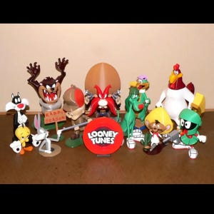 May include: A collection of Looney Tunes character figurines, including Taz, Bugs Bunny, Sylvester, and Yosemite Sam. The figurines are brightly colored and stand on a brown surface. A red Looney Tunes logo is also present.