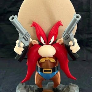 Puede incluir: Una figura impresa en 3D de Yosemite Sam, un personaje de dibujos animados, sosteniendo dos revólveres de juguete. La figura tiene un gran bigote rojo, un sombrero marrón y un atuendo azul y marrón. La figura está sobre una base rocosa.