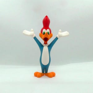 May include: A colorful Woody Woodpecker figurine. The cartoon character has a red head, orange beak and feet, and a blue body with a white chest. His arms are outstretched, and he has white gloves.