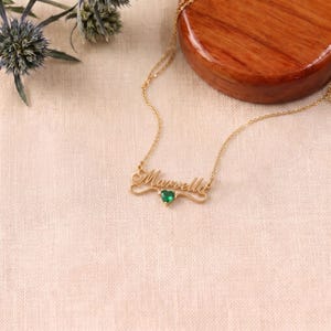 Può includere: Una collana color oro con una catenina sottile e un ciondolo con il nome Marvella in corsivo, impreziosito da una piccola pietra preziosa verde a forma di cuore posizionata esattamente sotto il centro del nome.