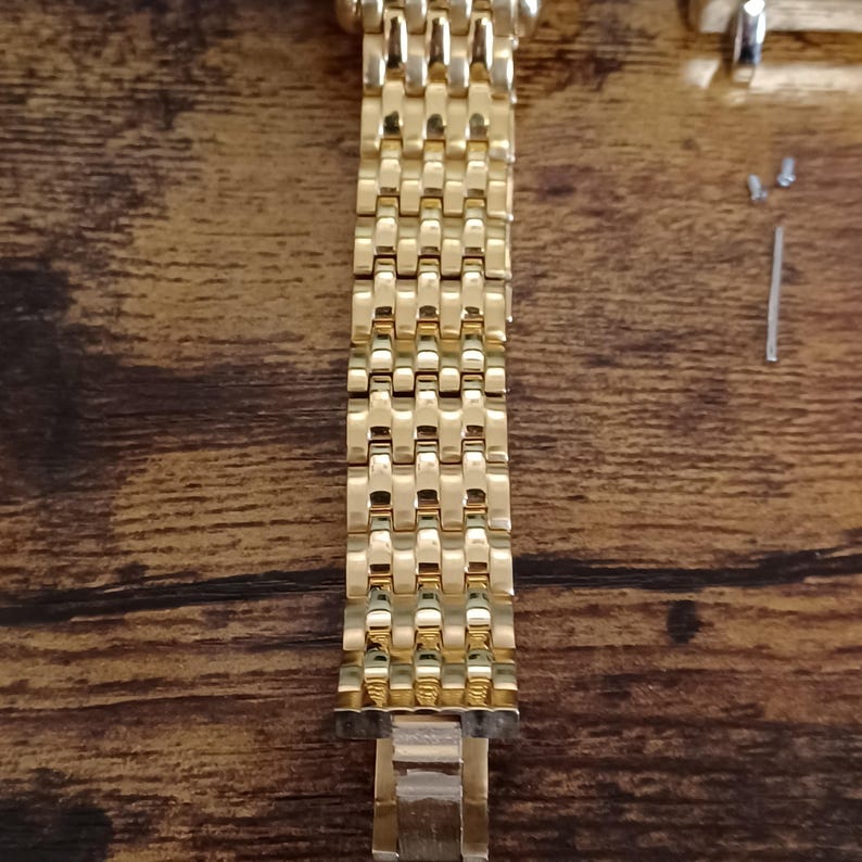 May include: A gold-colored watch band with a linked design. The band is made of metal and has a shiny finish. The band is lying on a wooden surface, with watch repair tools visible.