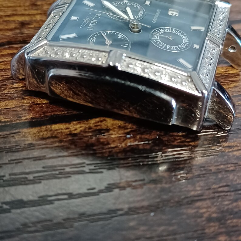 May include: A silver-toned Invicta watch with a rectangular face and a dark blue dial. The watch features multiple sub-dials, a date display, and a bezel encrusted with small, clear stones. The watch is set against a dark wooden surface.