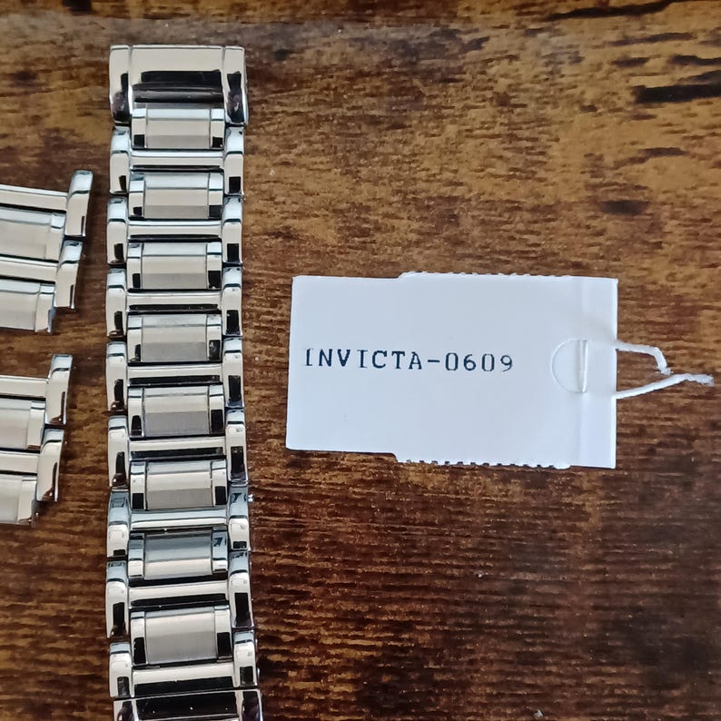 May include: A silver-toned watch band with rectangular links is displayed on a wooden surface. A white tag with the text "INVICTA-0609" is also present. The watch band is designed to be attached to a watch.
