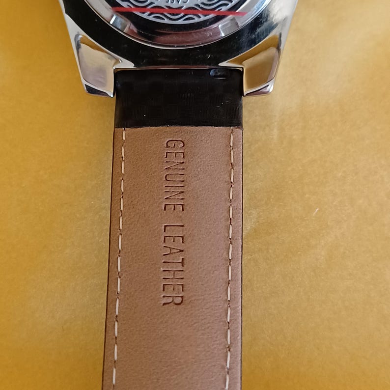 May include: Close-up of a watch with a black leather strap. The strap is tan with the words "GENUINE LEATHER" printed on it. The watch face is partially visible, with a silver case and a black band connecting to the strap.
