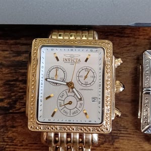 May include: Gold-toned Invicta chronograph watch with a square face and a gold band. The watch face is white with subdials and the days of the week. The bezel is encrusted with small, clear stones. The watch is water resistant to 100m.