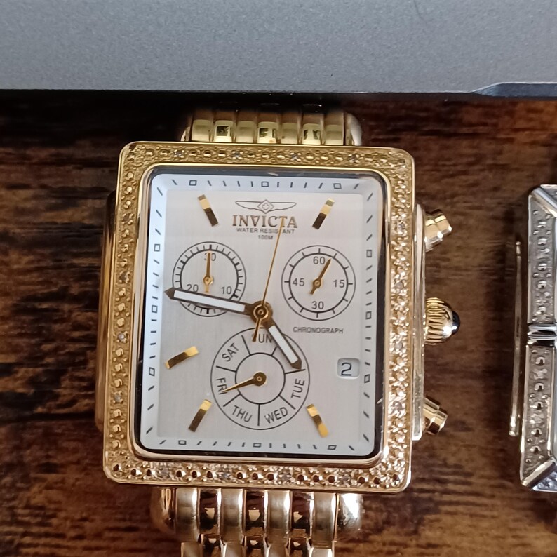 May include: Gold-toned Invicta chronograph watch with a square face and a gold band. The watch face is white with subdials and the days of the week. The bezel is encrusted with small, clear stones. The watch is water resistant to 100m.