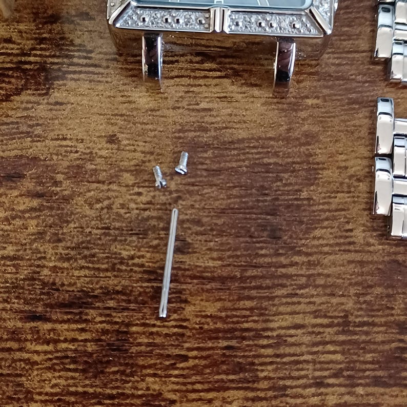 May include: Close-up of watch components on a wooden surface. The image shows a watch face with a silver band, small screws, and a silver pin. The watch face has a rectangular shape with a silver frame and decorative stones.