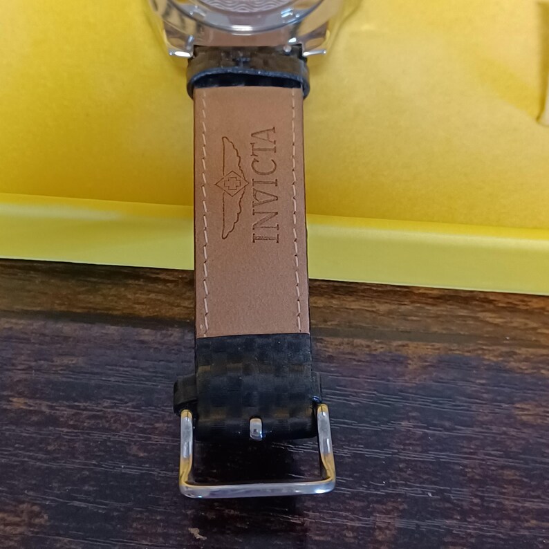 May include: Close-up of a watch with a black leather strap. The strap features the brand name "Invicta" embossed in the leather. The watch is set against a yellow background.