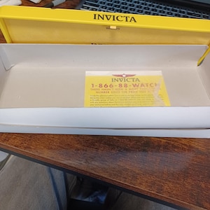 May include: An open yellow Invicta watch box with a white interior. A yellow card with the Invicta logo and the phone number 1-866-88-WATCH is inside the box. The Invicta logo and the word "Warranty" are printed on the box.