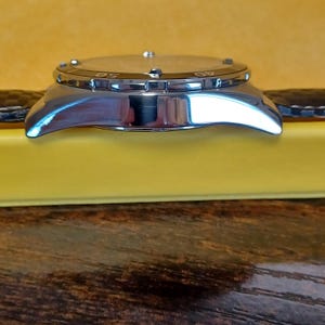 May include: A silver-toned wristwatch with a black leather band. The watch face is visible, with numbers marking the time. The watch is set on a yellow surface, with a wooden background.
