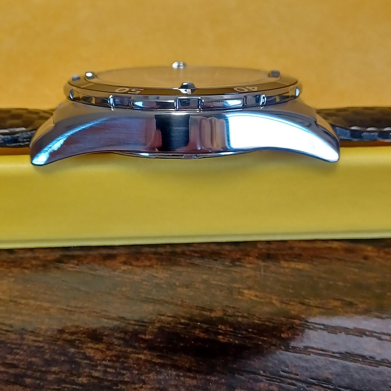 May include: A silver-toned wristwatch with a black leather band. The watch face is visible, with numbers marking the time. The watch is set on a yellow surface, with a wooden background.