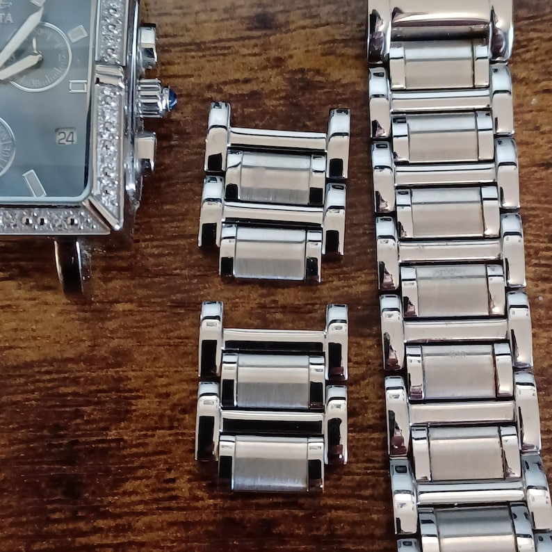 May include: A silver-toned rectangular watch with a diamond-studded bezel and a gray face, next to watch links and a watch band. The watch face displays the number 24. The watch and components are on a wooden surface.