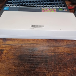May include: A white rectangular box with a barcode label, resting on a wooden surface. The box is positioned in front of a laptop keyboard. The label includes the text "J155894-00003-00241" and "IN3133".