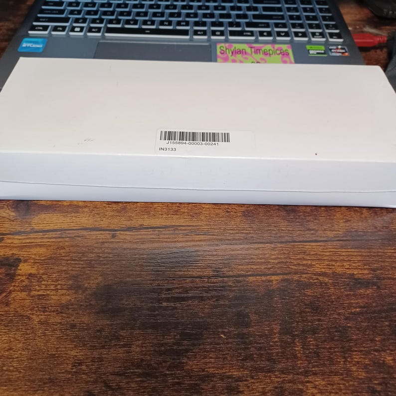 May include: A white rectangular box with a barcode label, resting on a wooden surface. The box is positioned in front of a laptop keyboard. The label includes the text "J155894-00003-00241" and "IN3133".