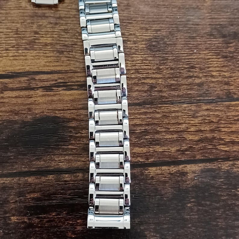 May include: A silver-colored watch band with a rectangular link design. The band is made of metal and has a polished, reflective surface. The watch band is shown against a brown wooden surface.