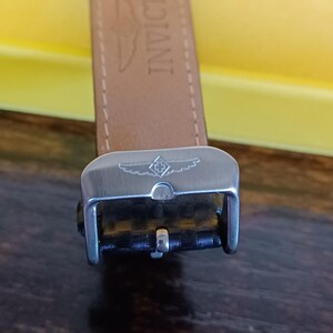 May include: Close-up of a watch strap and buckle. The strap is light brown leather with the word "INVICT" printed on it. The buckle is silver-colored metal with a textured black insert and a logo.