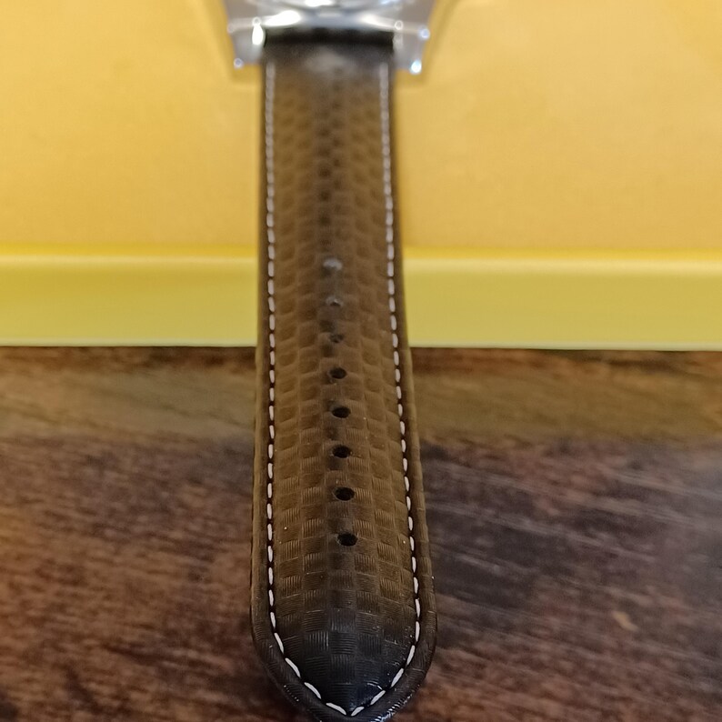 May include: Close-up of a watch with a dark brown leather strap. The strap has a textured pattern and white stitching along the edges. The watch face is partially visible at the top.