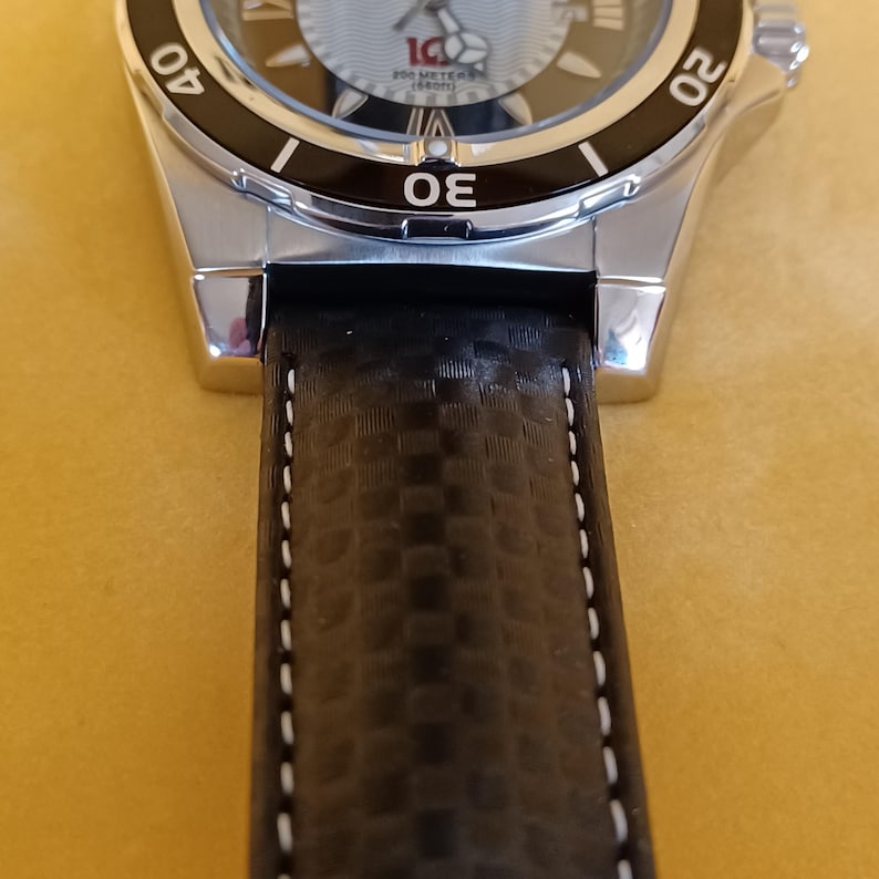 May include: Close-up of a silver wristwatch with a black leather band. The watch face has a light blue background with black and silver accents. The band is textured with white stitching.