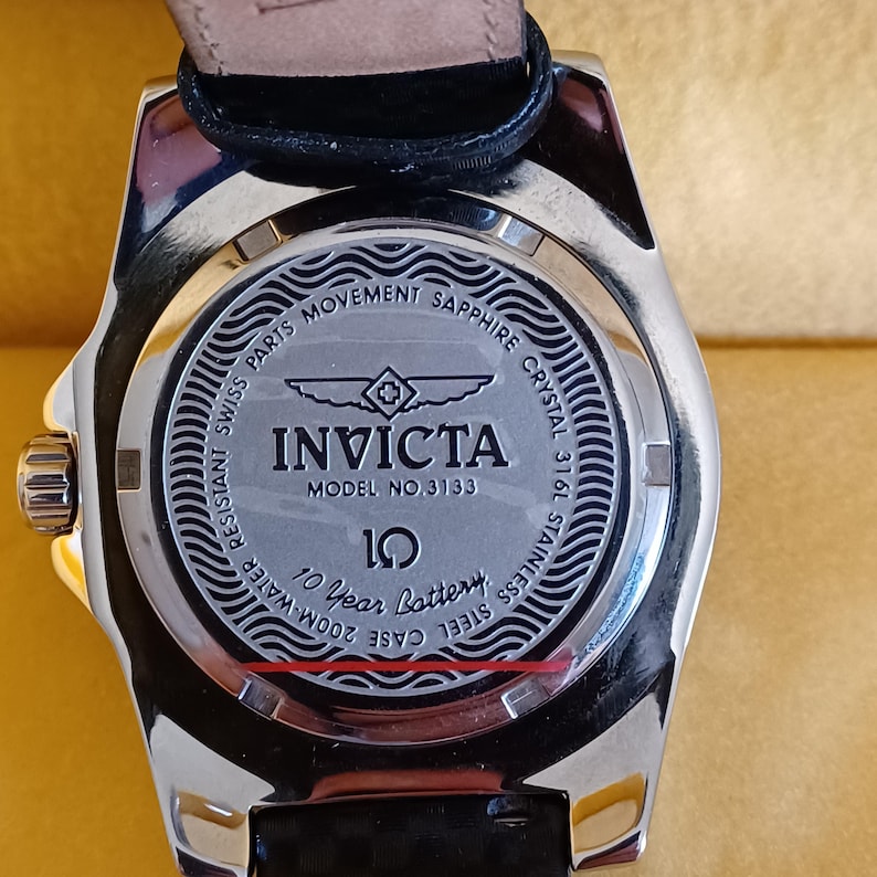 May include: Close-up of the back of an Invicta watch. The watch has a silver stainless steel case and a black leather strap. The watch face has the Invicta logo and text that reads "Model No. 3133", "Sapphire Crystal", and "10 Year Battery".