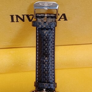 May include: Close-up of a black watch strap with white stitching and a silver buckle. The strap has a carbon fiber pattern. The word "Invicta" is visible on the background.