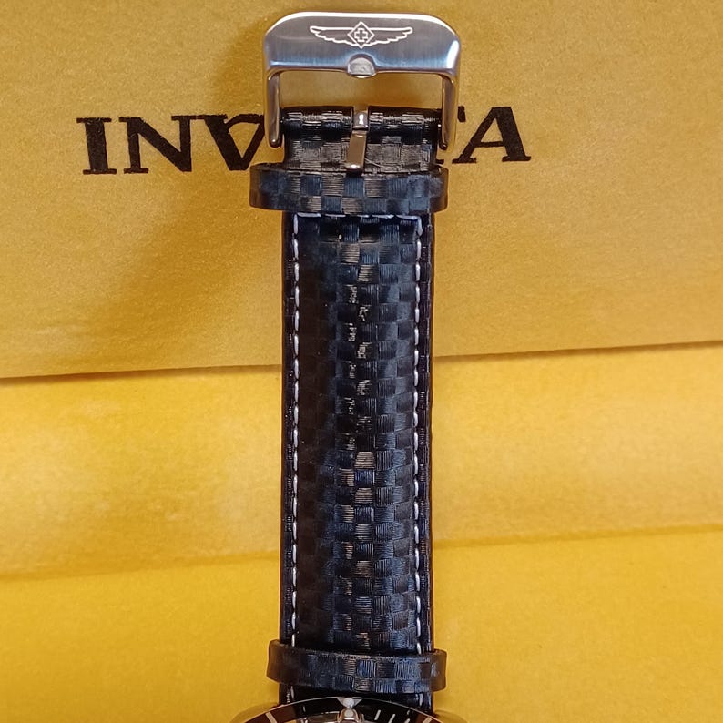 May include: Close-up of a black watch strap with white stitching and a silver buckle. The strap has a carbon fiber pattern. The word "Invicta" is visible on the background.