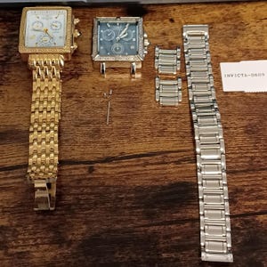 May include: Two square-faced Invicta watches, one gold-toned with a matching band, the other silver-toned with a blue face. Additional watch components and a silver band are also present. The watches have multiple dials and a textured bezel.