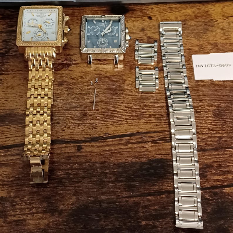 May include: Two square-faced Invicta watches, one gold-toned with a matching band, the other silver-toned with a blue face. Additional watch components and a silver band are also present. The watches have multiple dials and a textured bezel.