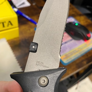 May include: Close-up of a silver and black folding knife. The blade is partially serrated and has the text "DELTA 6213 Taiwan" etched on it. The handle is black with two visible screws.