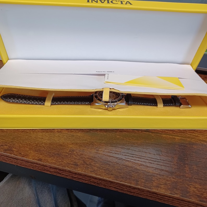 May include: An Invicta watch in a yellow box. The watch has a black leather band and a silver-colored face. The watch is secured in the box with yellow straps. The box is open, revealing the watch and a white interior.