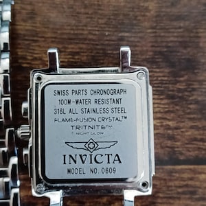 May include: Close-up of a silver Invicta watch face. The watch face has the text "SWISS PARTS CHRONOGRAPH", "100M-WATER RESISTANT", and "316L ALL STAINLESS STEEL". The watch also features the text "FLAME-FUSION CRYSTAL", "TRITNITE NIGHT GLOW", and "INVICTA MODEL NO. 0609".