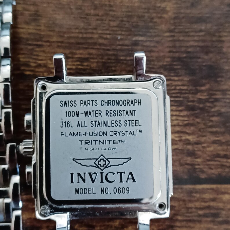 May include: Close-up of a silver Invicta watch face. The watch face has the text "SWISS PARTS CHRONOGRAPH", "100M-WATER RESISTANT", and "316L ALL STAINLESS STEEL". The watch also features the text "FLAME-FUSION CRYSTAL", "TRITNITE NIGHT GLOW", and "INVICTA MODEL NO. 0609".
