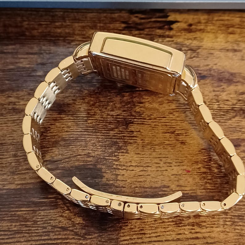 May include: A gold-toned wristwatch with a rectangular face and a linked band. The watch is displayed on a wooden surface. The band is partially unclasped, showing the inner workings of the clasp. The watch appears to be new.