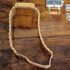 May include: A gold-toned rectangular wristwatch with a matching link bracelet. The watch is partially open, revealing the inner workings. The background is a dark wooden surface, with a second watch and small parts visible.