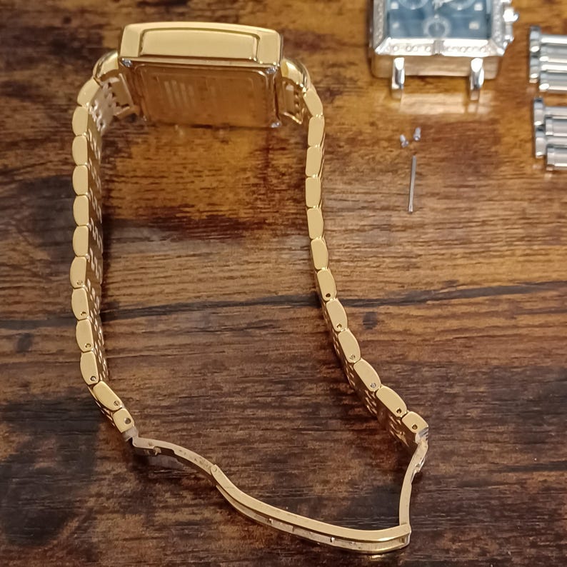 May include: A gold-toned rectangular wristwatch with a matching link bracelet. The watch is partially open, revealing the inner workings. The background is a dark wooden surface, with a second watch and small parts visible.