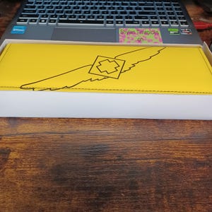 May include: A yellow rectangular wallet with a black wing design and a geometric shape in the center. The wallet is in a white box, resting on a wooden surface. The text "Shylan Timepieces on" is visible on a sticker.