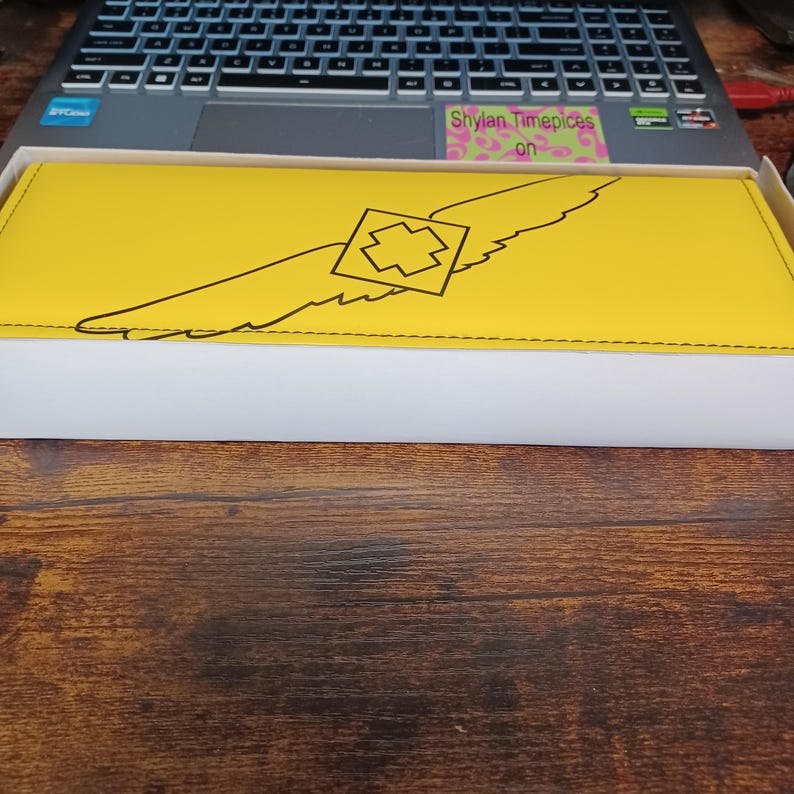 May include: A yellow rectangular wallet with a black wing design and a geometric shape in the center. The wallet is in a white box, resting on a wooden surface. The text "Shylan Timepieces on" is visible on a sticker.