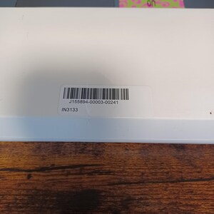 May include: A close-up of a white box with a barcode sticker. The sticker has the text "J155894-00003-00241" and "IN3133". The box is on a wooden surface.