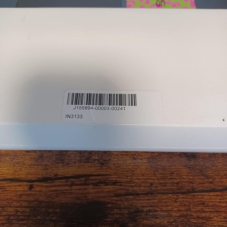 May include: A close-up of a white box with a barcode sticker. The sticker has the text "J155894-00003-00241" and "IN3133". The box is on a wooden surface.